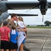 A military servicemember in flight suit is reuniting with his family on an airfield tarmac, sharing a kiss with a woman who is holding a baby. Two children stand nearby, and a handmade welcome home sign reading 'Daddy' is visible. A large military turboprop aircraft, consistent with a C-130 variant, is parked directly behind them.