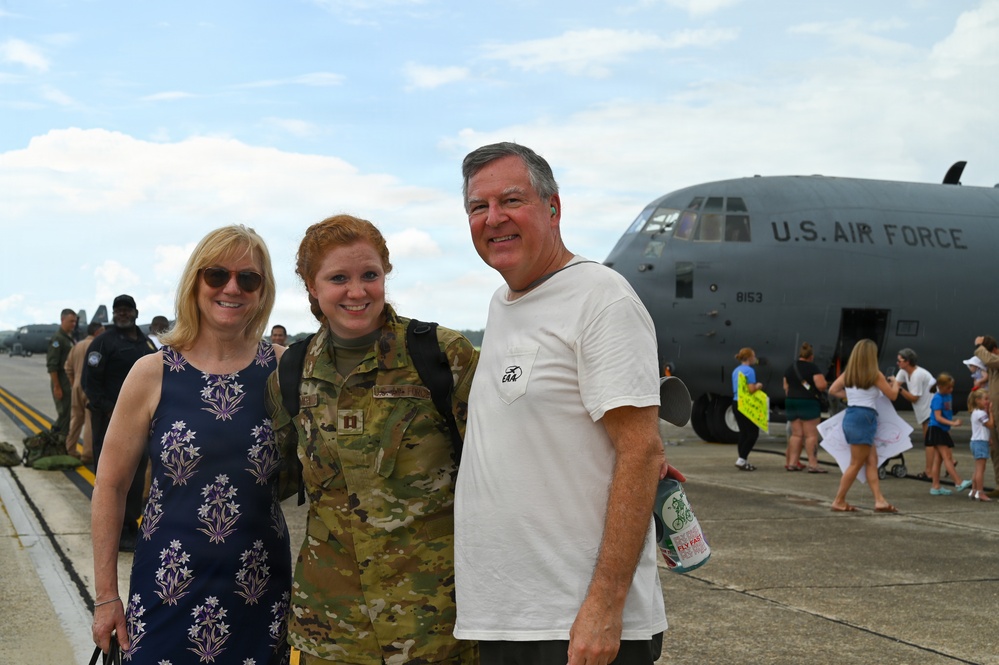 A U.S. Air Force service member in camouflage uniform is reuniting with two civilians, likely family members, on an airfield tarmac. A U.S. Air Force C-130 aircraft (tail number 8153) is visible in the background. The scene appears to be a military homecoming event with other families visible in the background.
