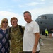 A U.S. Air Force service member in camouflage uniform is reuniting with two civilians, likely family members, on an airfield tarmac. A U.S. Air Force C-130 aircraft (tail number 8153) is visible in the background. The scene appears to be a military homecoming event with other families visible in the background.