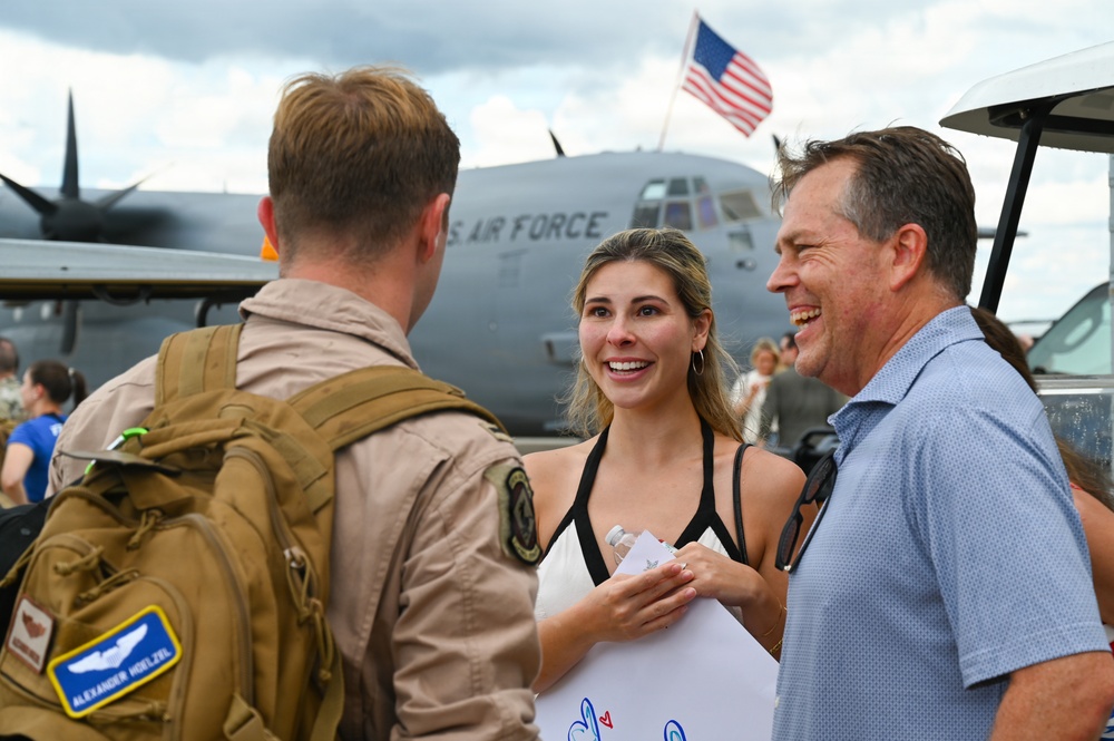 A U.S. Air Force service member in tan flight suit and tactical backpack is greeted by two civilians — a young woman and an older man — on an airfield tarmac. The scene appears to be a military homecoming or welcome event, with a large C-130 Hercules aircraft bearing 'U.S. AIR FORCE' markings visible in the background. An American flag flies above the aircraft, and other people can be seen gathered in the background.