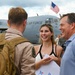 A U.S. Air Force service member in tan flight suit and tactical backpack is greeted by two civilians — a young woman and an older man — on an airfield tarmac. The scene appears to be a military homecoming or welcome event, with a large C-130 Hercules aircraft bearing 'U.S. AIR FORCE' markings visible in the background. An American flag flies above the aircraft, and other people can be seen gathered in the background.