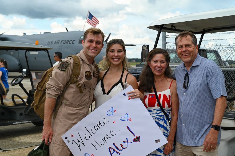 A U.S. Air Force service member named Alex is being welcomed home at a military airbase by what appears to be family members. The group of four people are smiling and posing together, with one person holding a handmade 'Welcome Home Alex!' sign. A U.S. Air Force aircraft is visible in the background along with an American flag.