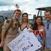 A U.S. Air Force service member named Alex is being welcomed home at a military airbase by what appears to be family members. The group of four people are smiling and posing together, with one person holding a handmade 'Welcome Home Alex!' sign. A U.S. Air Force aircraft is visible in the background along with an American flag.