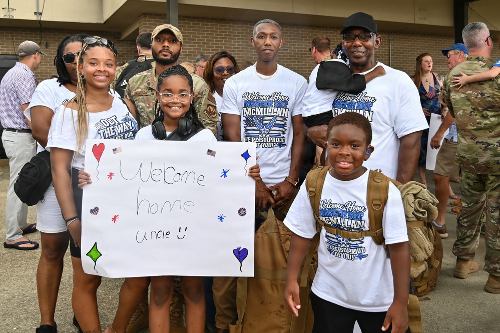 A family group is gathered at a military homecoming event, welcoming back a service member identified as 'Master Sgt. McMillan' based on the custom t-shirts worn by multiple family members. A young girl holds a handmade sign reading 'Welcome home Uncle' decorated with hearts and stars. The scene takes place outdoors near a brick building with other families and military personnel visible in the background.