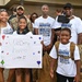 A family group is gathered at a military homecoming event, welcoming back a service member identified as 'Master Sgt. McMillan' based on the custom t-shirts worn by multiple family members. A young girl holds a handmade sign reading 'Welcome home Uncle' decorated with hearts and stars. The scene takes place outdoors near a brick building with other families and military personnel visible in the background.