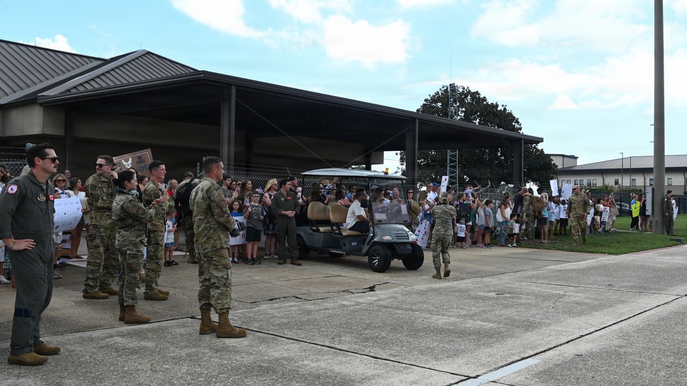 A military welcome home or greeting ceremony is taking place at what appears to be a U.S. Air Force base, evidenced by a visible Air Force emblem on a building. Military personnel in camouflage uniforms and at least one flight suit are gathered near a golf cart, while a large crowd of civilians including children hold signs and wave in celebration. The event appears to be a homecoming or deployment return ceremony.