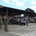 A military welcome home or greeting ceremony is taking place at what appears to be a U.S. Air Force base, evidenced by a visible Air Force emblem on a building. Military personnel in camouflage uniforms and at least one flight suit are gathered near a golf cart, while a large crowd of civilians including children hold signs and wave in celebration. The event appears to be a homecoming or deployment return ceremony.