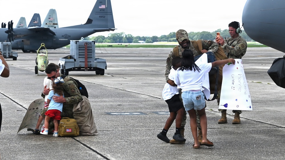 Military personnel are reuniting with family members on an airfield tarmac, likely following a deployment return. Two service members in camouflage uniforms are embracing children, while a third documents the moment on a phone. A handmade welcome sign reading 'Home Uncle U' is visible, indicating a planned homecoming celebration.