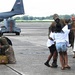 Military personnel are reuniting with family members on an airfield tarmac, likely following a deployment return. Two service members in camouflage uniforms are embracing children, while a third documents the moment on a phone. A handmade welcome sign reading 'Home Uncle U' is visible, indicating a planned homecoming celebration.