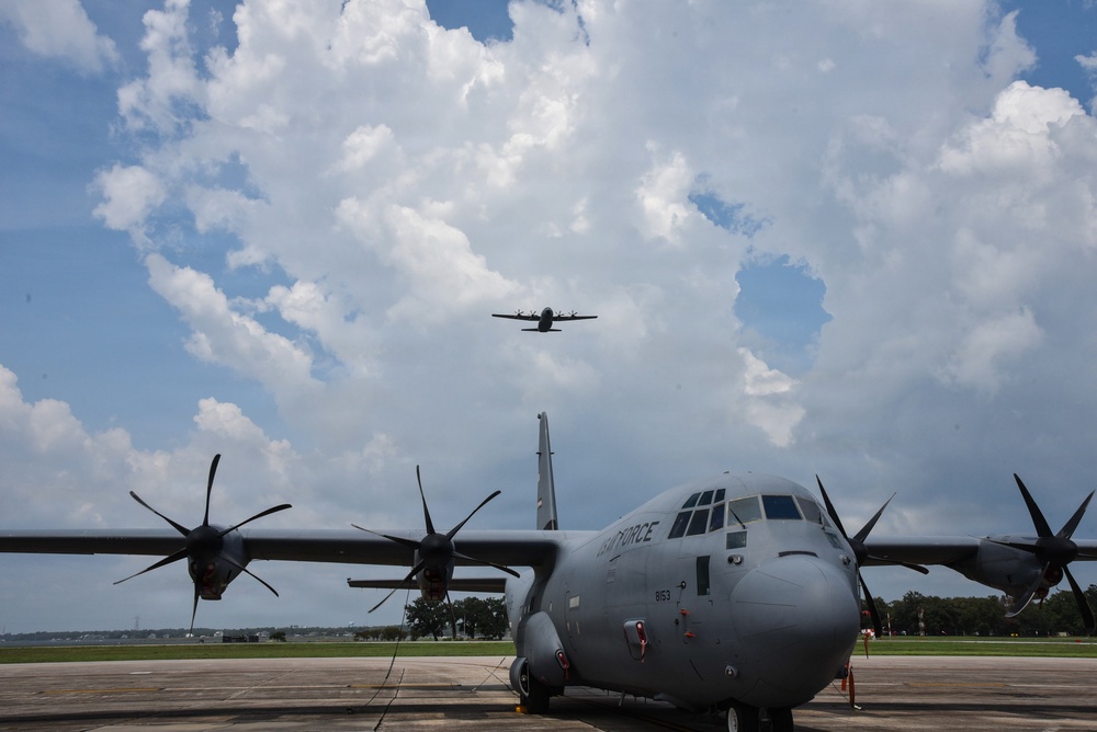 A U.S. Air Force C-130J Super Hercules turboprop military transport aircraft is parked on a flight line tarmac, with a second similar aircraft visible in flight overhead. The foreground aircraft bears the tail number 8153 and 'AIR FORCE' markings on the fuselage. The scene appears to be at a military air base under partly cloudy summer skies.