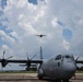 A U.S. Air Force C-130J Super Hercules turboprop military transport aircraft is parked on a flight line tarmac, with a second similar aircraft visible in flight overhead. The foreground aircraft bears the tail number 8153 and 'AIR FORCE' markings on the fuselage. The scene appears to be at a military air base under partly cloudy summer skies.