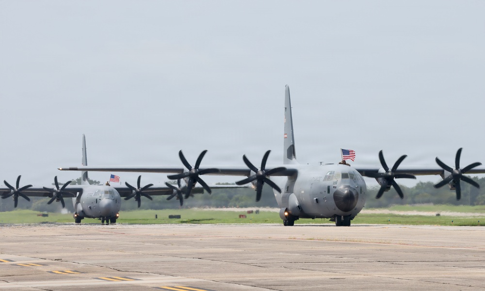 Two U.S. military C-130 Hercules turboprop transport aircraft are taxiing on a runway or tarmac at a military airfield. Both aircraft are painted in standard gray military livery and display American flags, suggesting a ceremonial or homecoming event. The propellers on both aircraft are spinning, indicating active engine operation.