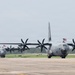 Two U.S. military C-130 Hercules turboprop transport aircraft are taxiing on a runway or tarmac at a military airfield. Both aircraft are painted in standard gray military livery and display American flags, suggesting a ceremonial or homecoming event. The propellers on both aircraft are spinning, indicating active engine operation.