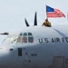 A U.S. Air Force C-130 Hercules military transport aircraft, tail number 8159, is taxiing on a runway. A person in military uniform is visible emerging from a top hatch of the aircraft, holding an American flag. The scene appears to be a ceremonial or homecoming arrival event.