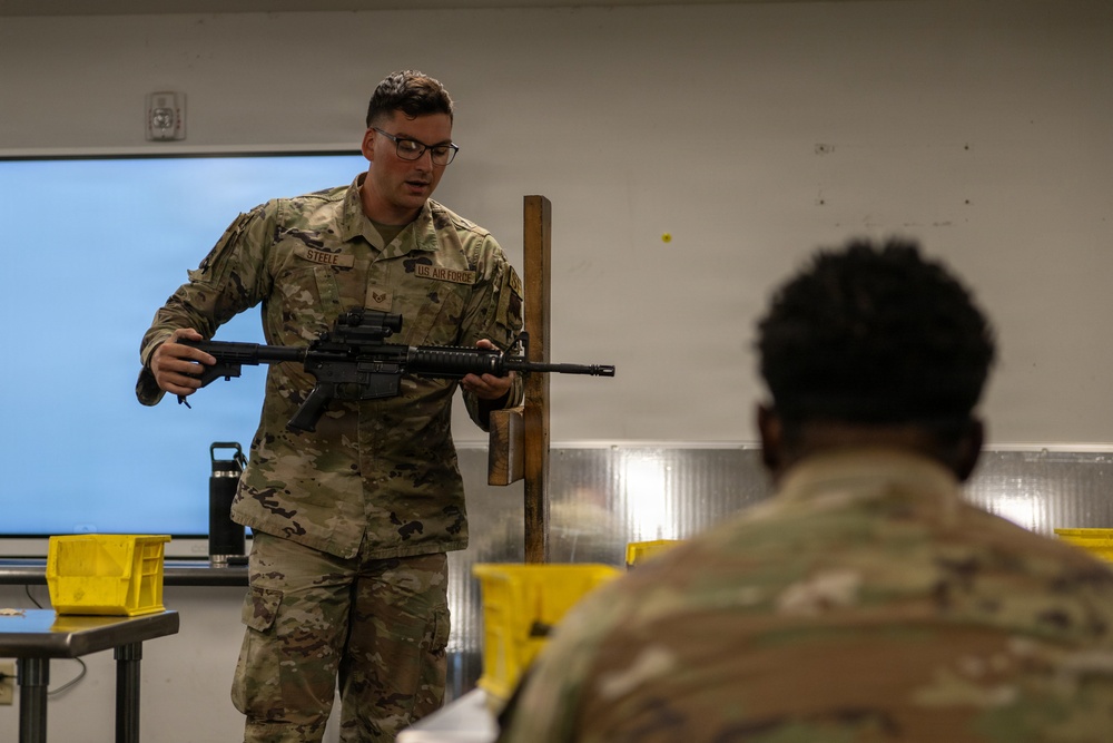 A U.S. Air Force instructor, identified by name tape as 'Steele,' conducts a weapons familiarization or training class in an indoor classroom setting. He is holding and presenting an M4/AR-style carbine rifle to at least one seated student in military uniform. The setting appears to be a formal military training environment with tables, yellow bins, and a whiteboard visible.