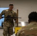 A U.S. Air Force instructor, identified by name tape as 'Steele,' conducts a weapons familiarization or training class in an indoor classroom setting. He is holding and presenting an M4/AR-style carbine rifle to at least one seated student in military uniform. The setting appears to be a formal military training environment with tables, yellow bins, and a whiteboard visible.