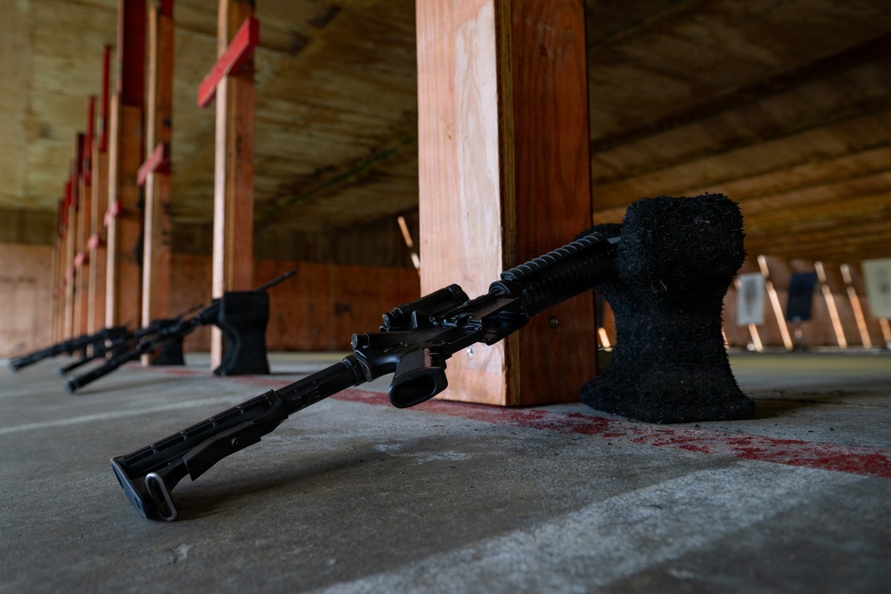 The image shows multiple rifles laid on the concrete floor of what appears to be an outdoor shooting range. The weapons are resting against black sandbag-style supports between wooden divider posts. Paper targets are visible downrange in the background.