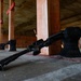 The image shows multiple rifles laid on the concrete floor of what appears to be an outdoor shooting range. The weapons are resting against black sandbag-style supports between wooden divider posts. Paper targets are visible downrange in the background.