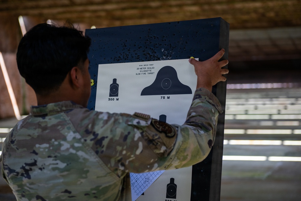 A soldier in MultiCam/OCP camouflage uniform is posting or examining a paper shooting target on a dark backing board at a firing range. The target is a M19A1 Series Target featuring 25-meter scaled silhouette slow fire targets representing 300M and 75M distances. The backing board shows numerous bullet holes from previous firing sessions.