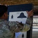 A soldier in MultiCam/OCP camouflage uniform is posting or examining a paper shooting target on a dark backing board at a firing range. The target is a M19A1 Series Target featuring 25-meter scaled silhouette slow fire targets representing 300M and 75M distances. The backing board shows numerous bullet holes from previous firing sessions.
