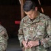 Two U.S. Air Force personnel in OCP (Operational Camouflage Pattern) uniforms are visible inside what appears to be a wooden structure, likely a shooting range or training facility. The foreground subject, identified by name tape as 'RASK,' holds the rank of First Lieutenant (single silver bar) and is handling a firearm magazine. A second airman is partially visible on the left side of the frame.