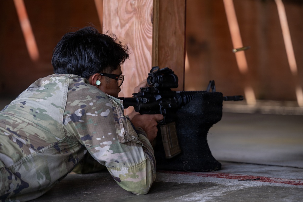 A person wearing military-pattern camouflage (OCP/Multicam) is in a prone shooting position, aiming a rifle equipped with an optic sight around a wooden barrier. The shooter is wearing glasses and has a distinctive ear gauge/plug visible. The setting appears to be an indoor or covered shooting range with a concrete floor.