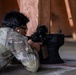 A person wearing military-pattern camouflage (OCP/Multicam) is in a prone shooting position, aiming a rifle equipped with an optic sight around a wooden barrier. The shooter is wearing glasses and has a distinctive ear gauge/plug visible. The setting appears to be an indoor or covered shooting range with a concrete floor.