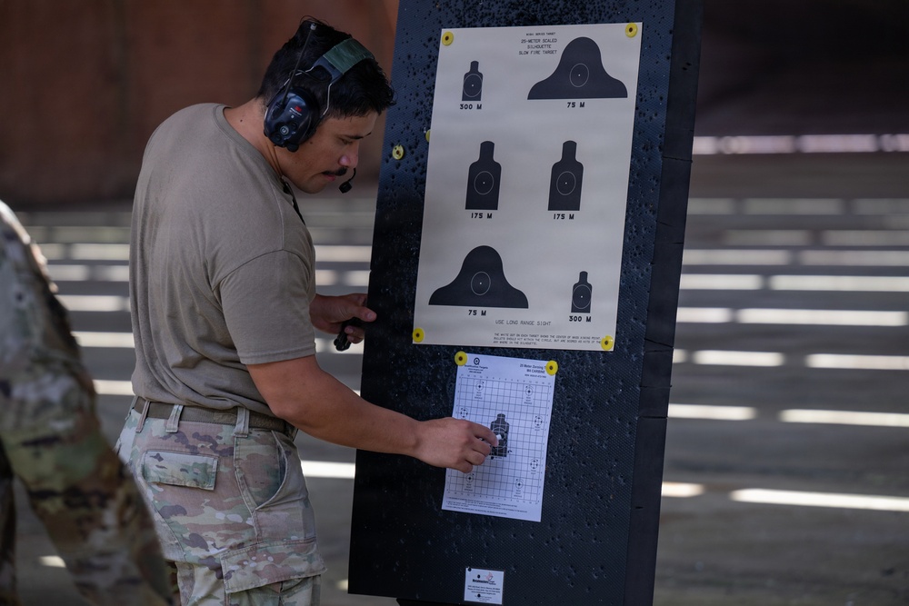 A military or law enforcement personnel wearing hearing protection and a tan t-shirt with multicam pants is examining a shooting target board at a firing range. The board displays a standard M16/M4 25-meter scaled silhouette qualification target chart showing distances of 75M, 175M, and 300M. Below the reference chart, a zeroing or grouping target sheet with bullet hole patterns is attached for evaluation.