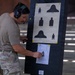 A military or law enforcement personnel wearing hearing protection and a tan t-shirt with multicam pants is examining a shooting target board at a firing range. The board displays a standard M16/M4 25-meter scaled silhouette qualification target chart showing distances of 75M, 175M, and 300M. Below the reference chart, a zeroing or grouping target sheet with bullet hole patterns is attached for evaluation.