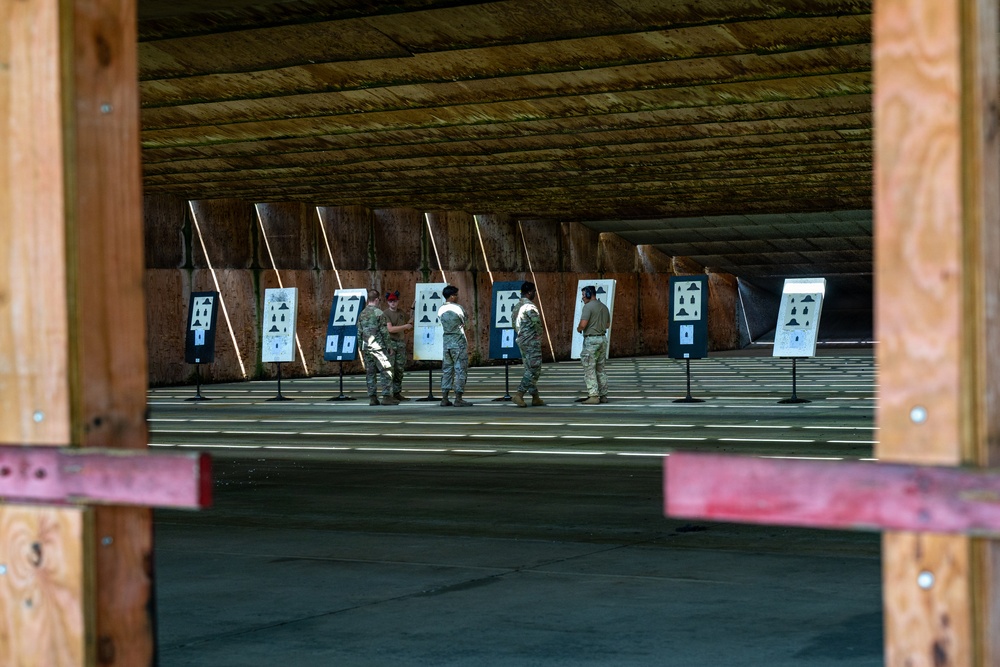 Military personnel in camouflage uniforms are gathered at an indoor shooting range, examining paper targets on stands. The range features a covered structure with a wooden ceiling and earthen/sand backstop berm. Approximately four to five individuals appear to be reviewing or scoring their targets after a shooting exercise.