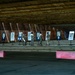 Military personnel in camouflage uniforms are gathered at an indoor shooting range, examining paper targets on stands. The range features a covered structure with a wooden ceiling and earthen/sand backstop berm. Approximately four to five individuals appear to be reviewing or scoring their targets after a shooting exercise.