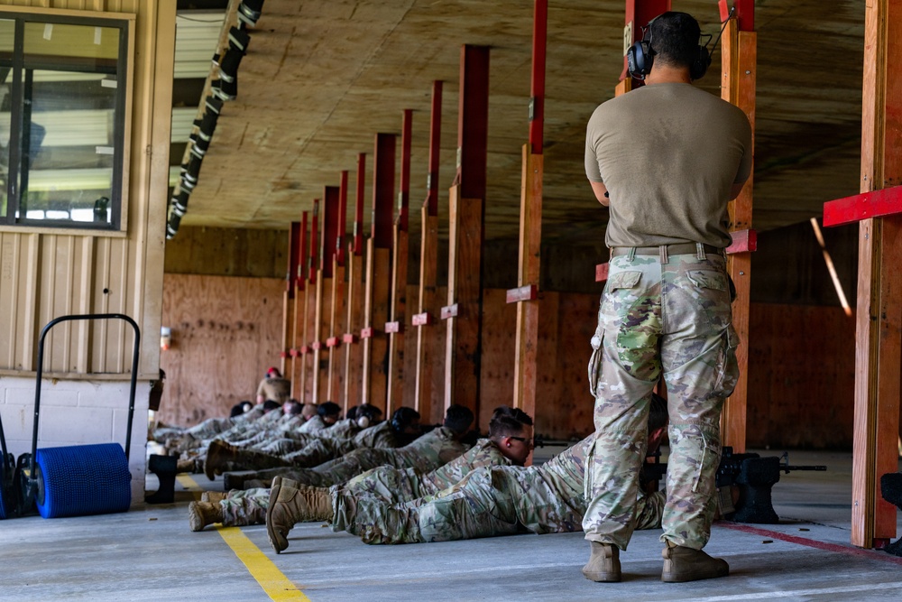 Military personnel are conducting a weapons qualification or marksmanship training exercise at an indoor shooting range. Multiple soldiers in OCP (Operational Camouflage Pattern) uniforms are in the prone position along a firing line, while a standing instructor or range safety officer observes from the rear. The facility appears to be a covered range with wooden target backer frames extending down the lane.