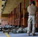 Military personnel are conducting a weapons qualification or marksmanship training exercise at an indoor shooting range. Multiple soldiers in OCP (Operational Camouflage Pattern) uniforms are in the prone position along a firing line, while a standing instructor or range safety officer observes from the rear. The facility appears to be a covered range with wooden target backer frames extending down the lane.