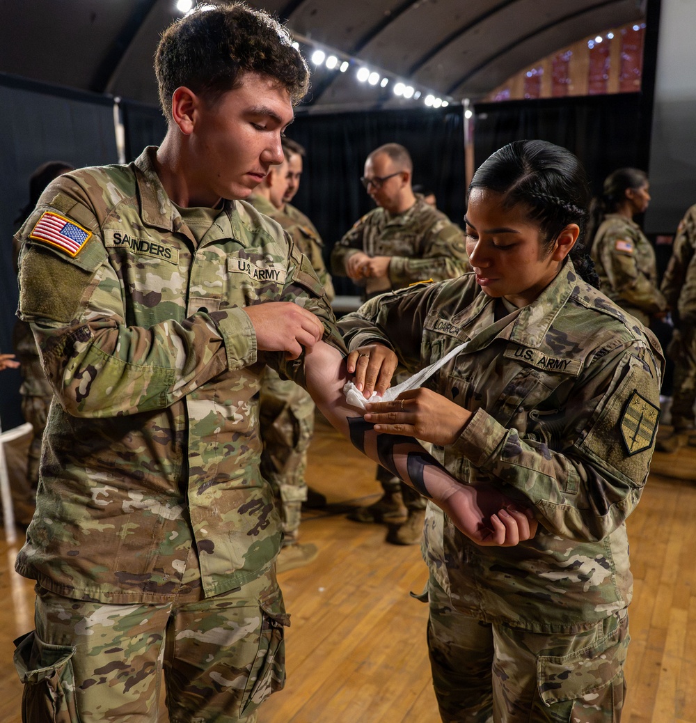 Two U.S. Army soldiers in OCP (Operational Camouflage Pattern) uniforms are engaged in what appears to be a medical or first aid procedure, with one soldier applying a bandage or swab to the other's arm. The setting appears to be an indoor facility with a wooden floor and overhead lighting, with additional soldiers visible in the background performing similar activities. The name tapes visible identify the soldiers as 'SAUNDERS' and 'LOARCA'.