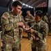 Two U.S. Army soldiers in OCP (Operational Camouflage Pattern) uniforms are engaged in what appears to be a medical or first aid procedure, with one soldier applying a bandage or swab to the other's arm. The setting appears to be an indoor facility with a wooden floor and overhead lighting, with additional soldiers visible in the background performing similar activities. The name tapes visible identify the soldiers as 'SAUNDERS' and 'LOARCA'.