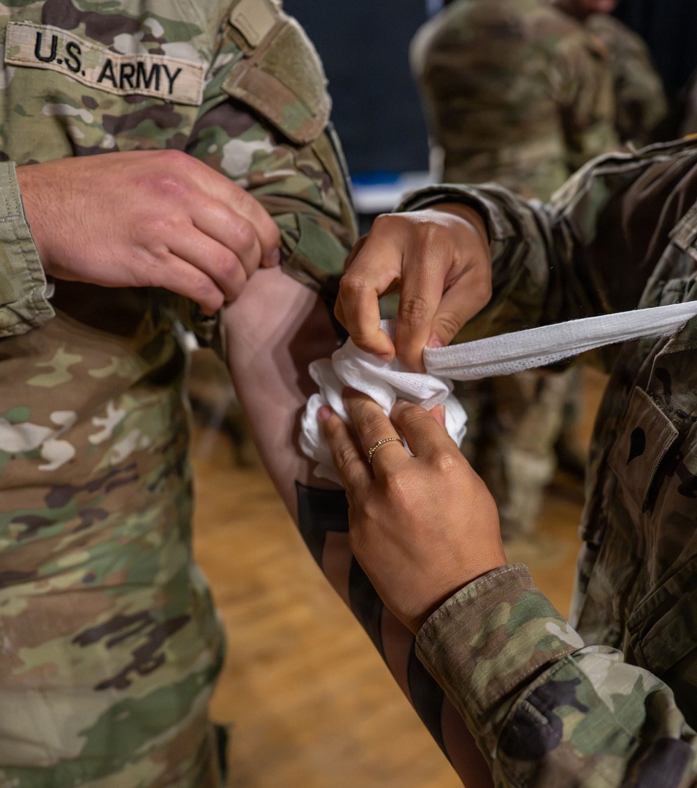 Two U.S. Army soldiers in OCP (Operational Camouflage Pattern) uniforms are practicing first aid bandaging techniques. One soldier is applying a white gauze/bandage wrap to the arm of another soldier, likely during a medical training exercise. The close-up shot focuses on the hands and the bandaging process.
