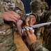 Two U.S. Army soldiers in OCP (Operational Camouflage Pattern) uniforms are practicing first aid bandaging techniques. One soldier is applying a white gauze/bandage wrap to the arm of another soldier, likely during a medical training exercise. The close-up shot focuses on the hands and the bandaging process.