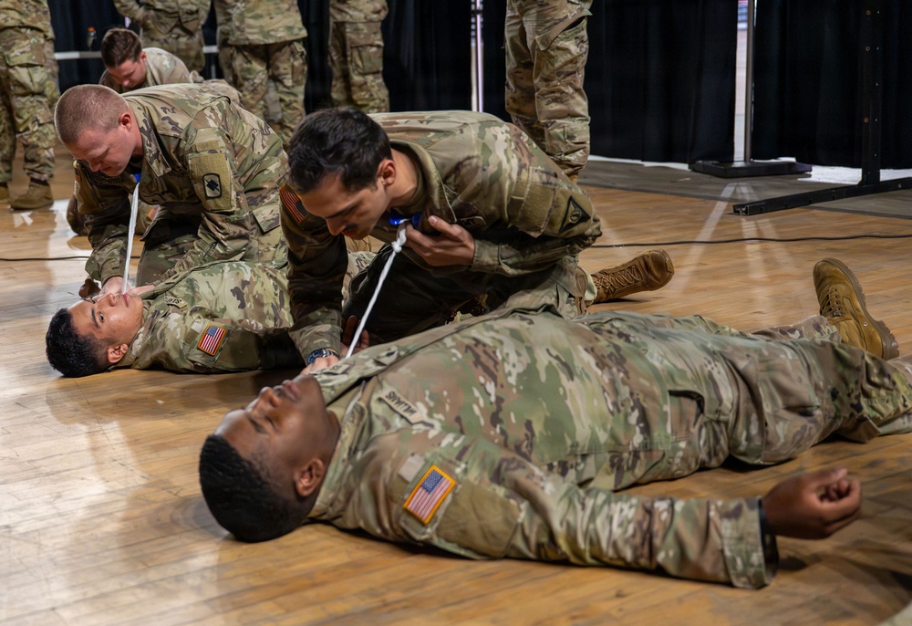Military personnel in US Army OCP (Operational Camouflage Pattern) uniforms are conducting a combat casualty care or first aid training exercise on a gymnasium floor. Two soldiers are lying on the ground acting as simulated casualties while others kneel over them performing medical procedures. A second group is visible in the background conducting a similar exercise.