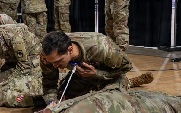 Military personnel in US Army OCP (Operational Camouflage Pattern) uniforms are conducting a combat casualty care or first aid training exercise on a gymnasium floor. Two soldiers are lying on the ground acting as simulated casualties while others kneel over them performing medical procedures. A second group is visible in the background conducting a similar exercise.