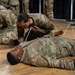 Military personnel in US Army OCP (Operational Camouflage Pattern) uniforms are conducting a combat casualty care or first aid training exercise on a gymnasium floor. Two soldiers are lying on the ground acting as simulated casualties while others kneel over them performing medical procedures. A second group is visible in the background conducting a similar exercise.