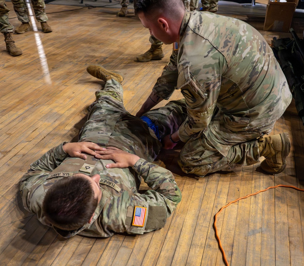 Two U.S. Army soldiers in OCP (Operational Camouflage Pattern) uniforms are engaged in what appears to be a combat casualty care or first aid training exercise on a wooden gymnasium floor. One soldier (name tape reads 'TURNER') is lying face-down acting as the simulated casualty, while a second soldier kneels beside him applying what appears to be a tourniquet or bandage to the midsection/hip area. Additional personnel in military uniforms are visible standing in the background.