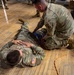 Two U.S. Army soldiers in OCP (Operational Camouflage Pattern) uniforms are engaged in what appears to be a combat casualty care or first aid training exercise on a wooden gymnasium floor. One soldier (name tape reads 'TURNER') is lying face-down acting as the simulated casualty, while a second soldier kneels beside him applying what appears to be a tourniquet or bandage to the midsection/hip area. Additional personnel in military uniforms are visible standing in the background.