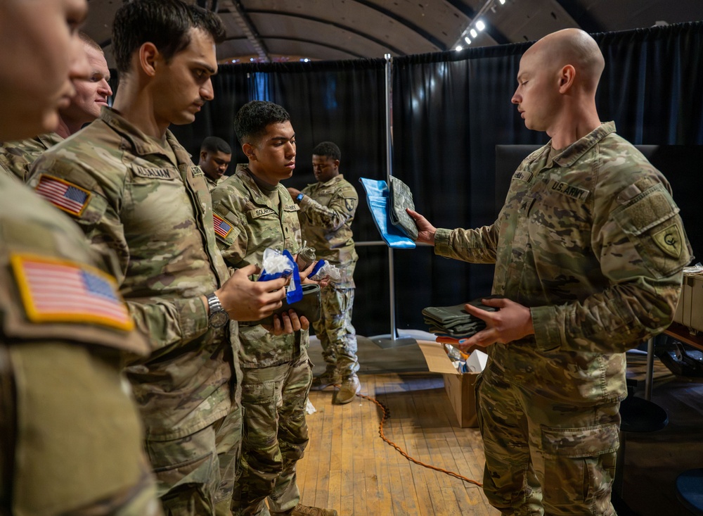 A group of U.S. Army soldiers in MultiCam/OCP camouflage uniforms are gathered indoors, appearing to receive or inspect equipment or supplies. One soldier on the right, identified by a 'U.S. ARMY' tape, is holding up a blue item and distributing materials from a cardboard box. The setting appears to be an indoor venue with black curtain backdrops and wooden flooring.