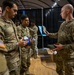 A group of U.S. Army soldiers in MultiCam/OCP camouflage uniforms are gathered indoors, appearing to receive or inspect equipment or supplies. One soldier on the right, identified by a 'U.S. ARMY' tape, is holding up a blue item and distributing materials from a cardboard box. The setting appears to be an indoor venue with black curtain backdrops and wooden flooring.