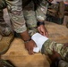 A soldier in OCP (Operational Camouflage Pattern) uniform is applying a white gauze bandage wrap to another soldier's forearm or wrist area. The setting appears to be an indoor training environment with a wooden floor, suggesting a medical training or first aid exercise. The soldier applying the bandage is wearing a black wristband and a tactical watch.