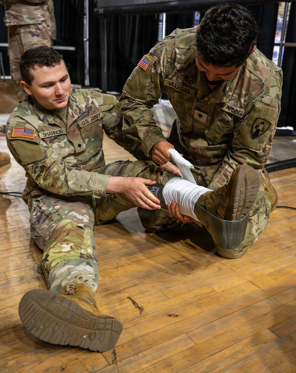 Two U.S. Army soldiers in OCP (Operational Camouflage Pattern) uniforms are engaged in a medical training exercise on a hardwood floor. Soldier 'VILLEGAS,' wearing a 1st Cavalry Division patch, is applying a white bandage/wrap to the lower leg or ankle of Soldier 'SHANNON,' who is seated on the floor. The setting appears to be an indoor training facility or gymnasium.