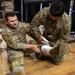 Two U.S. Army soldiers in OCP (Operational Camouflage Pattern) uniforms are engaged in a medical training exercise on a hardwood floor. Soldier 'VILLEGAS,' wearing a 1st Cavalry Division patch, is applying a white bandage/wrap to the lower leg or ankle of Soldier 'SHANNON,' who is seated on the floor. The setting appears to be an indoor training facility or gymnasium.
