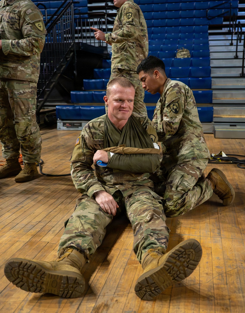 U.S. Army soldiers are conducting a combat casualty care or first aid training exercise inside a gymnasium or arena with blue bleacher seating. One soldier is seated on the hardwood floor with his arm in a sling/improvised splint, while a second soldier kneels behind him applying or adjusting the bandaging. The setting and uniforms indicate this is a structured military medical training scenario.