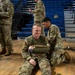 U.S. Army soldiers are conducting a combat casualty care or first aid training exercise inside a gymnasium or arena with blue bleacher seating. One soldier is seated on the hardwood floor with his arm in a sling/improvised splint, while a second soldier kneels behind him applying or adjusting the bandaging. The setting and uniforms indicate this is a structured military medical training scenario.