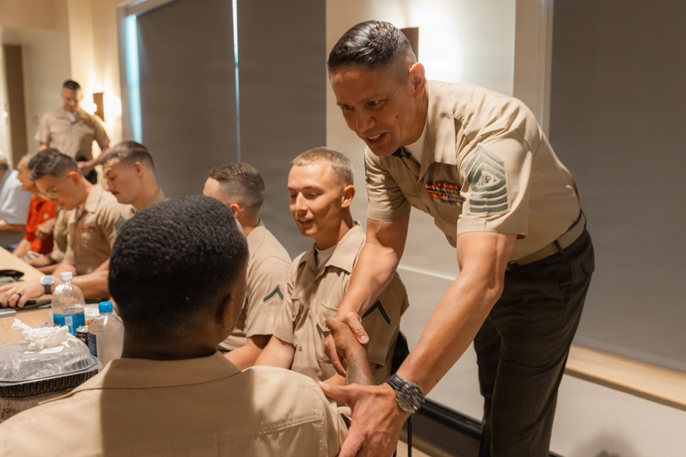 A senior U.S. Marine Corps non-commissioned officer, identifiable by his rank insignia and service ribbons, is leaning forward to shake hands with a seated Marine. The setting appears to be an indoor meeting or briefing room with multiple Marines seated at a table. The interaction suggests a formal or ceremonial greeting between a senior leader and junior enlisted personnel.